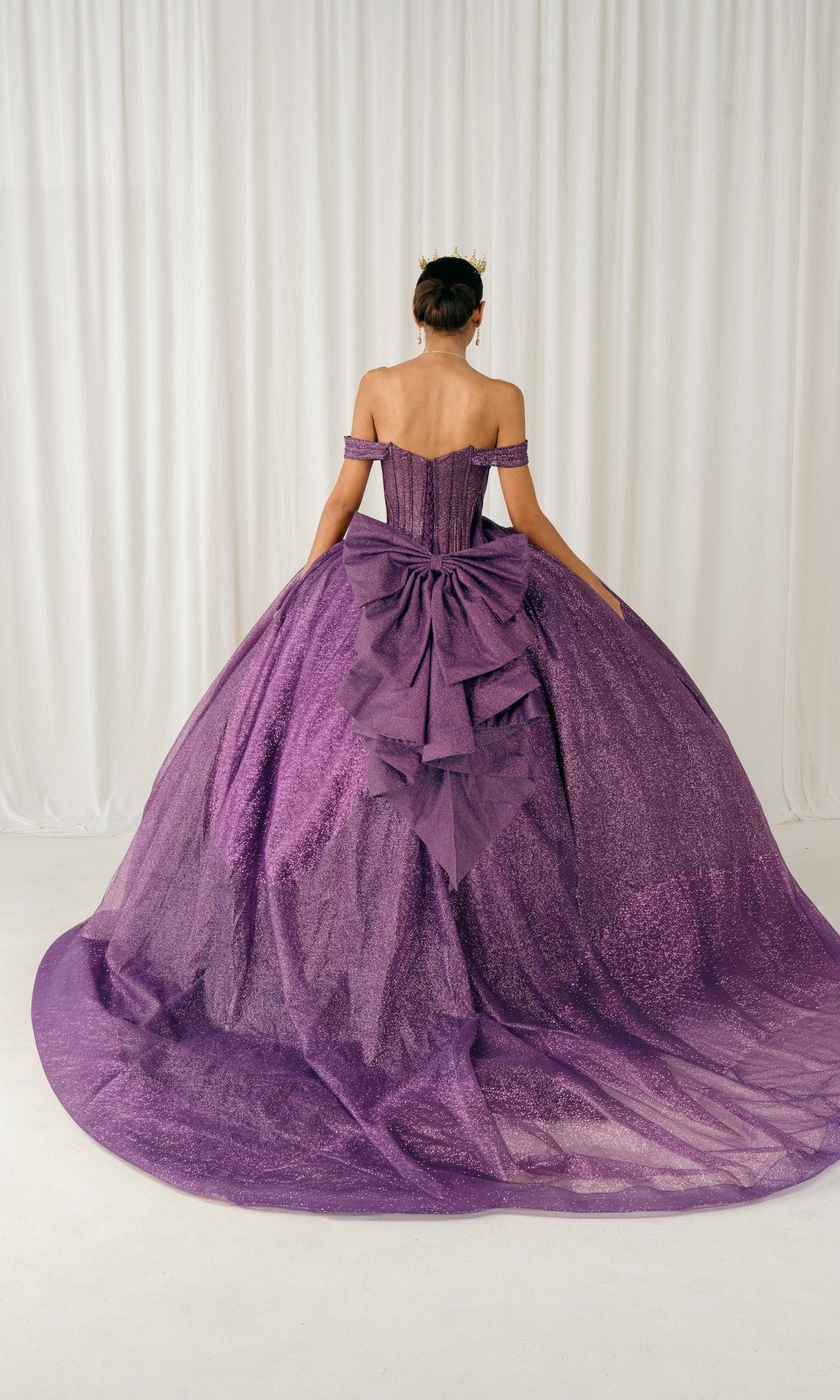 A woman faces away, wearing the Quinceanera Dress 8460JQ by Cinderella Couture—an off-the-shoulder purple gown with sparkles, a large bow, and a layered tulle skirt—standing before a white curtain backdrop.